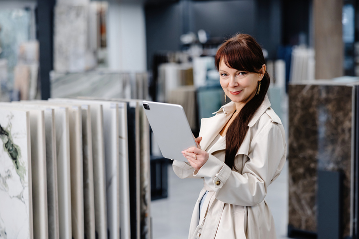 A woman in a light trench coat holding a tablet stands among vertical stone slab displays in a high-end showroom.