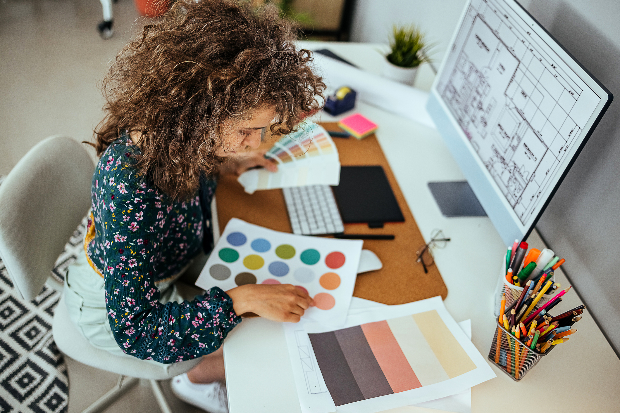 Interior designer working at her desk, reviewing color palettes and architectural floor plans on a computer, surrounded by design tools and stationery.